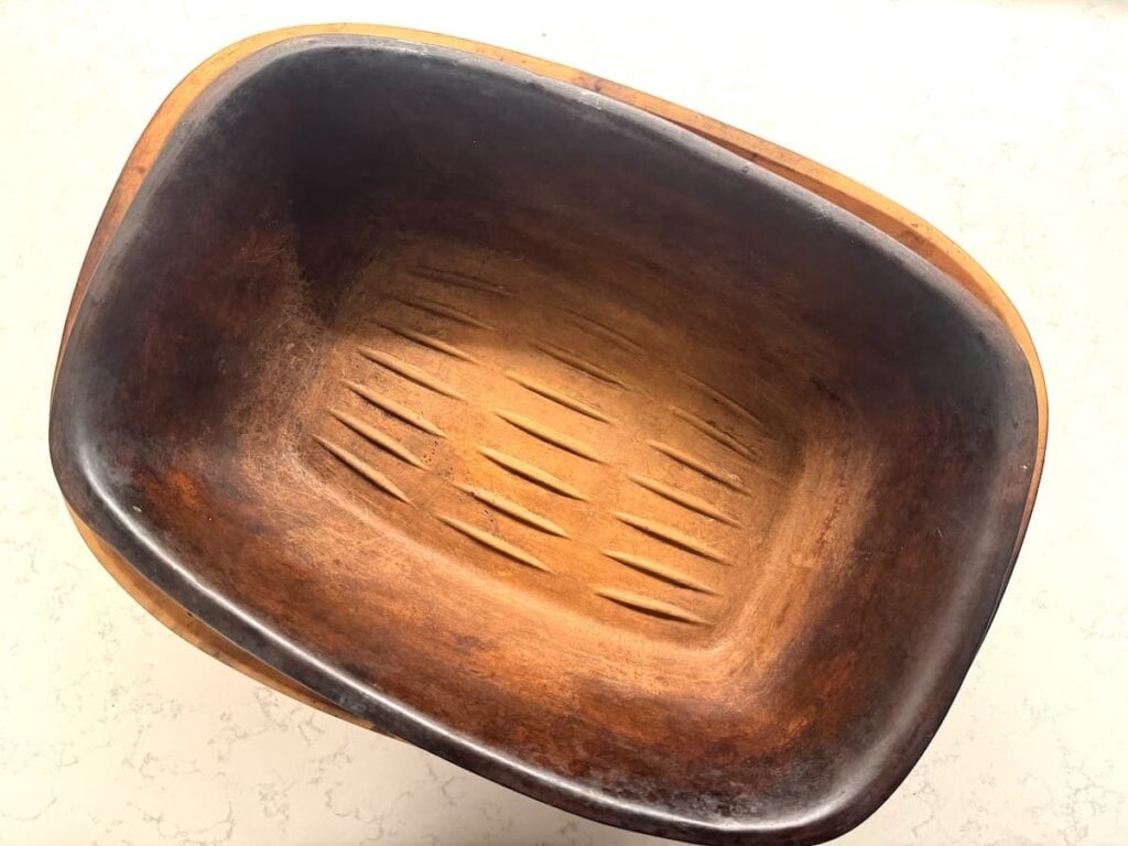A rectangular wooden bowl with a dark, worn interior and carved grooves on the bottom sits on a light-colored, textured surface, evoking the rustic charm of traditional clay pot chicken dishes.