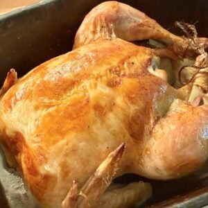 A whole roasted chicken with crispy, golden-brown skin sits in a dark roasting pan, reminiscent of classic clay pot chicken, with its legs tied together and onions visible underneath.