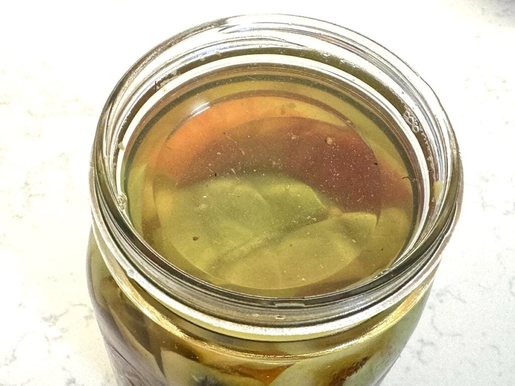 A close-up of a glass jar filled with homemade apple cider vinegar pickling liquid and large vegetable pieces, viewed from above on a light-colored surface.