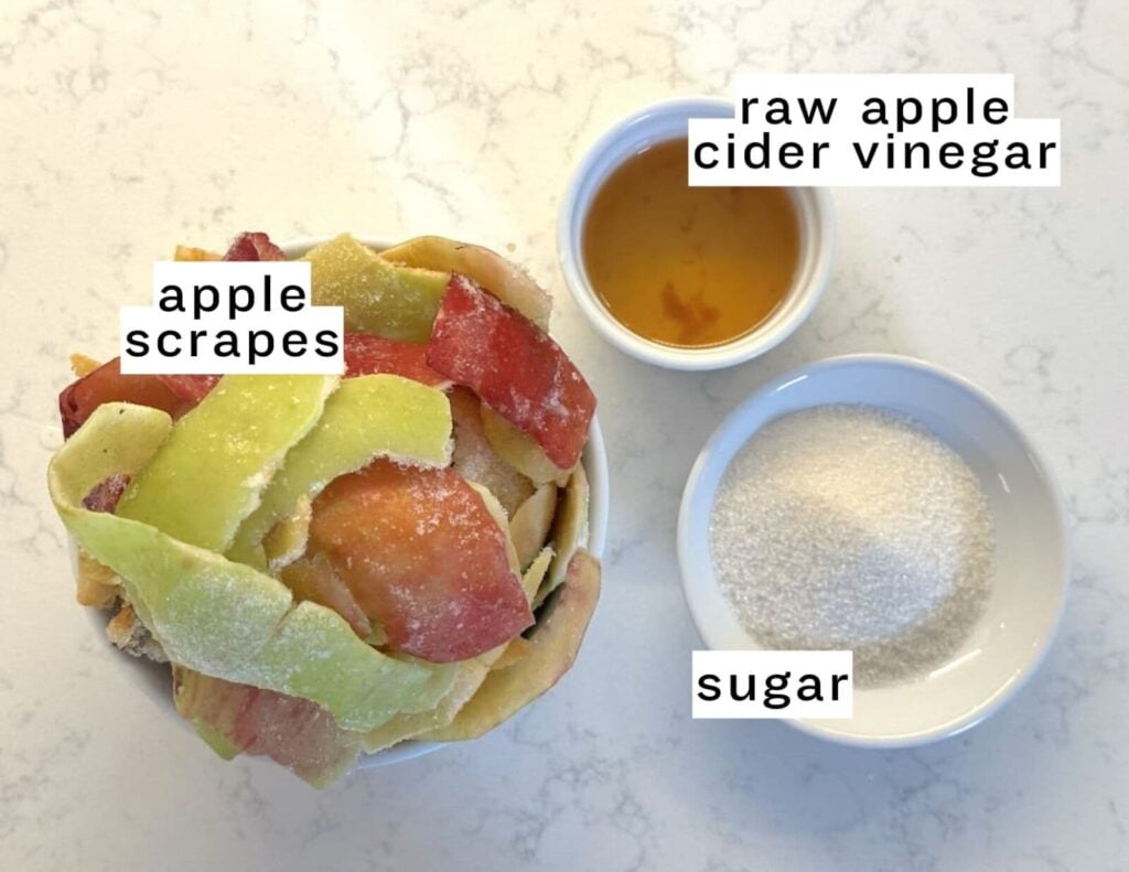 A bowl of apple scraps, a small bowl of raw homemade apple cider vinegar, and a small bowl of sugar are arranged on a white surface. Each ingredient is labeled.