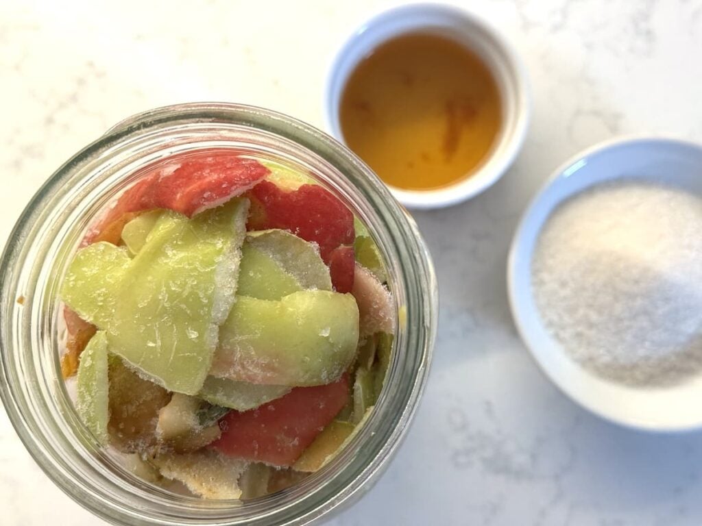 A top view of a glass jar filled with colorful apple peels for homemade apple cider vinegar, next to a small bowl of granulated sugar and a bowl of apple cider vinegar