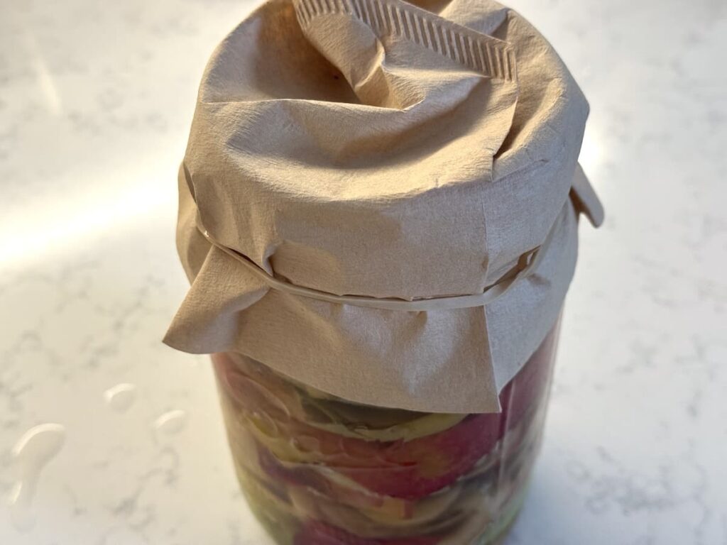 A glass jar filled with sliced vegetables and homemade apple cider vinegar, covered with brown paper and secured with a rubber band, sits on a light-colored, speckled countertop.