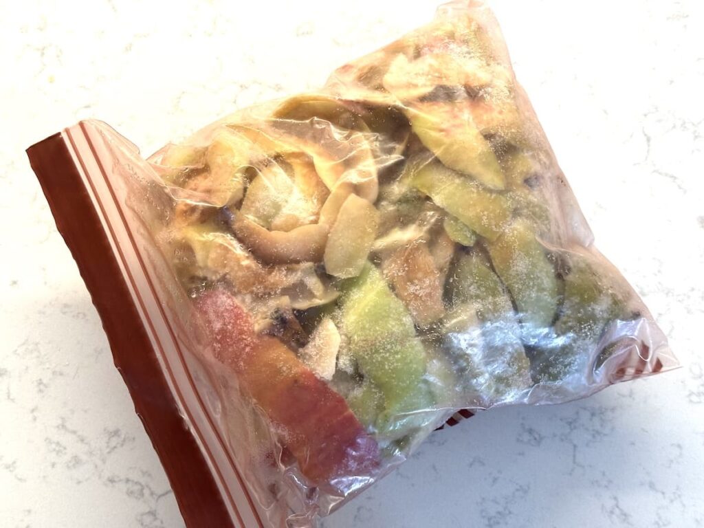 A clear plastic zipper bag filled with frozen apple peels - ideal for homemade apple cider vinegar - shows a mix of red, green, and yellow colors, resting on a white countertop.