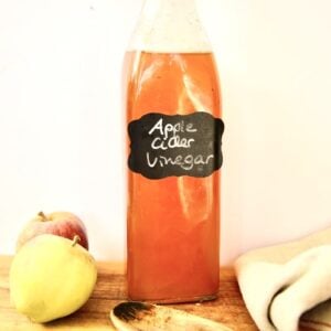A glass bottle labeled homemade apple cider vinegar sits on a wooden surface next to two apples, a wooden spoon, and a folded beige cloth with red stripes. The background is plain and light-colored.