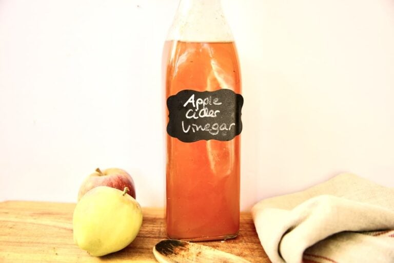 A glass bottle labeled homemade apple cider vinegar sits on a wooden surface next to two apples, a wooden spoon, and a folded beige cloth with red stripes. The background is plain and light-colored.