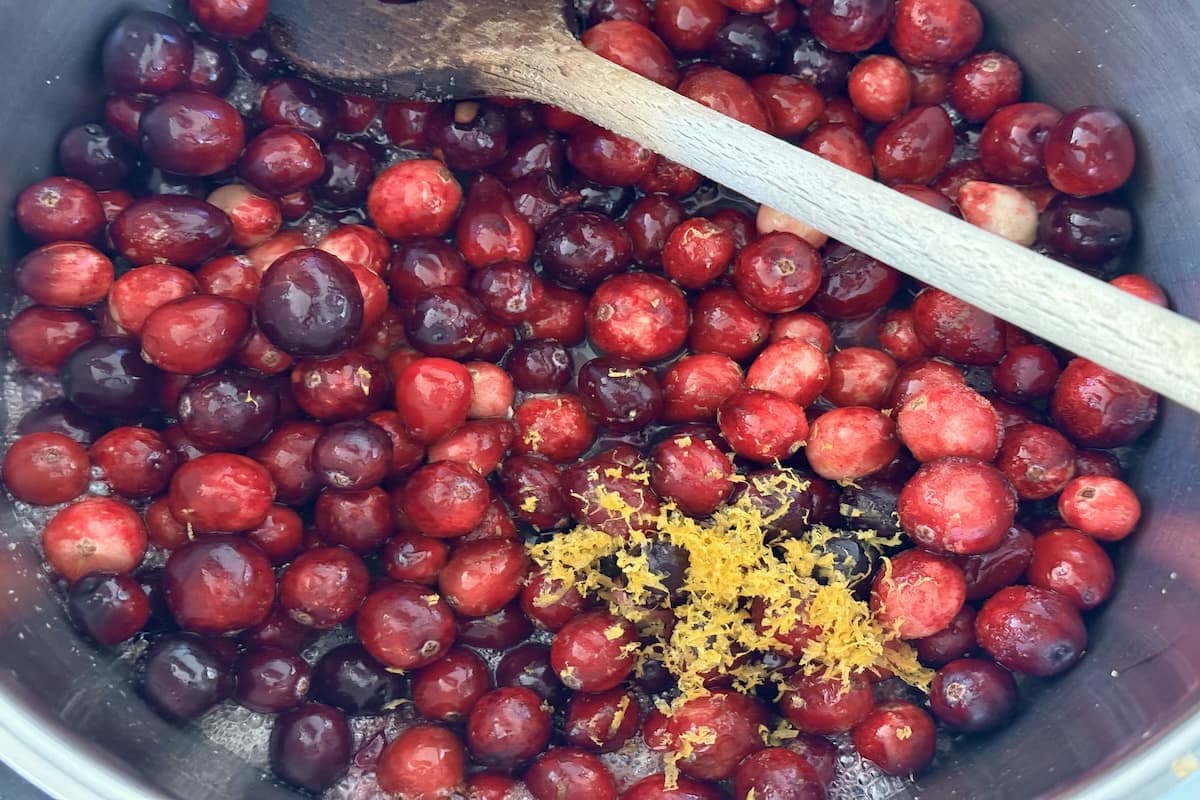 Whole cranberries and fresh orange zest in a saucepan with a wooden spoon.