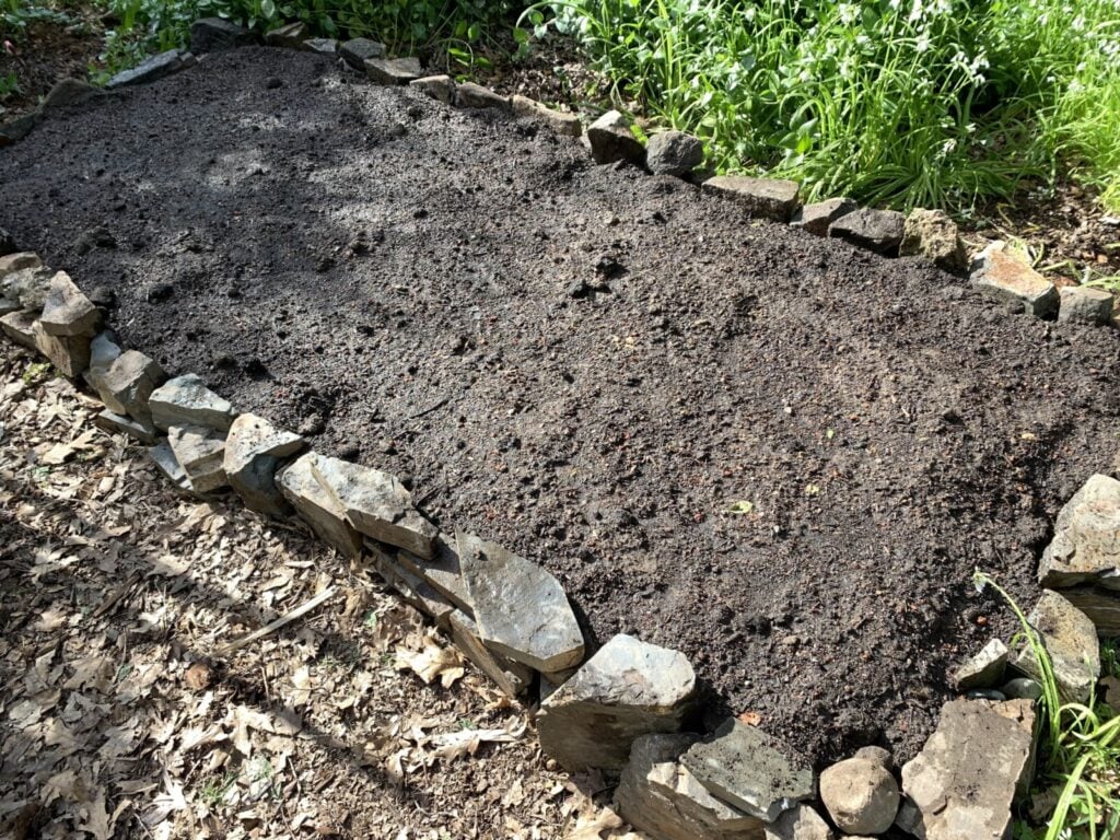 A rectangular raised garden bed bordered with uneven stones is filled with dark, freshly turned soil. Green plants and leaves surround the bed, suggesting a natural, outdoor setting.