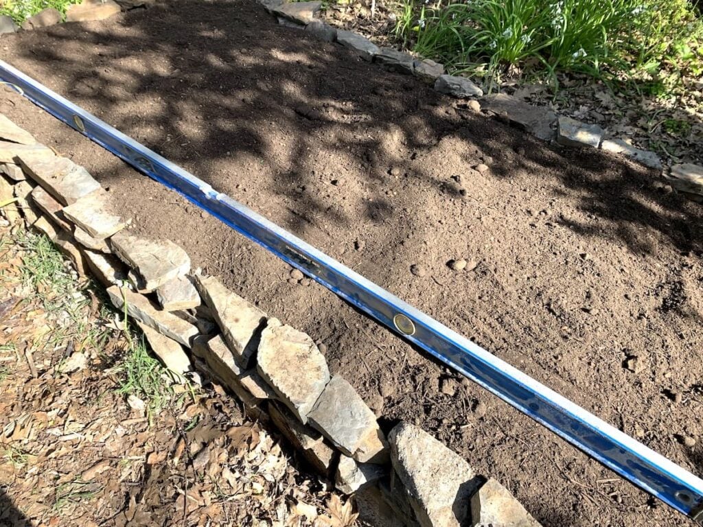 A stacked stone raised garden bed filled with soil, with a blue level tool resting diagonally across the edge to check evenness. Sunlight casts shadows over the raised garden beds area.