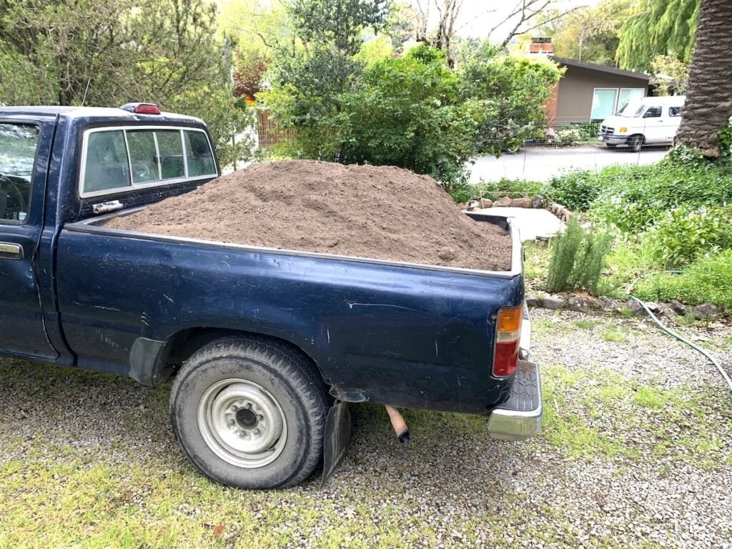 A blue pickup truck parked on gravel, its bed filled to the top with soil for raised garden beds. Green trees, shrubs, and a residential street are visible in the background.