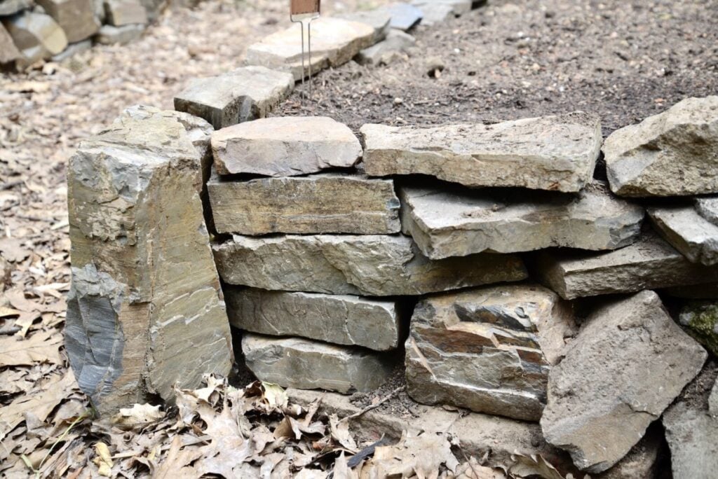 A close-up of a small stone retaining wall with irregularly stacked rocks borders raised garden beds filled with soil, while dry leaves are scattered on the ground in front of the wall.