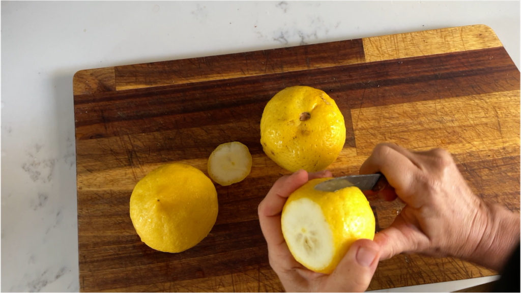 Two yellow lemons, one partially peeled with a small knife, rest on a wooden cutting board.