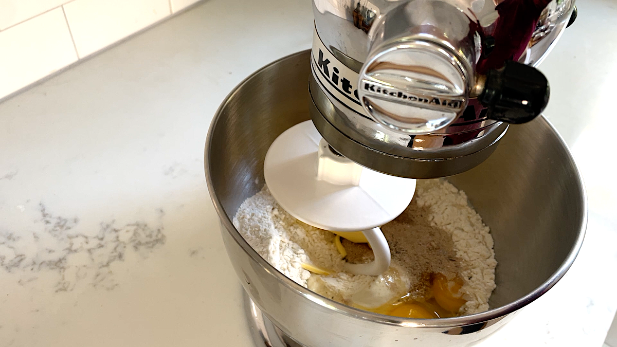 A stand mixer on a kitchen countertop contains flour, eggs, and other bread baking ingredients.