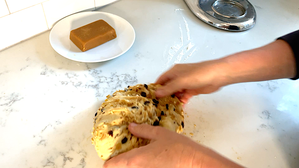 Hands kneading raisins, candied lemon peel, and chopped nuts into Christmas stollen dough.