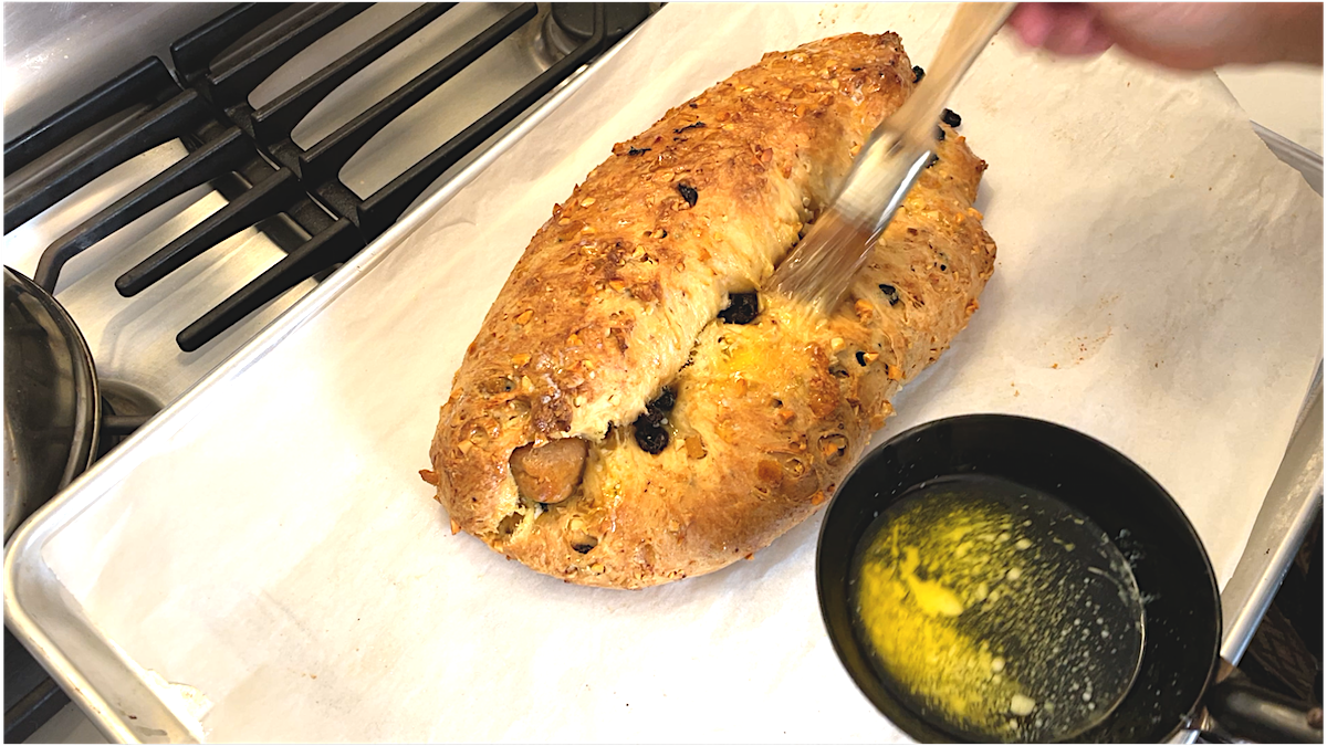 A hand brushes melted butter over a freshly baked Christmas Stollen, studded with raisins, on a parchment-lined baking tray. A black cup of melted butter sits nearby on the tray.