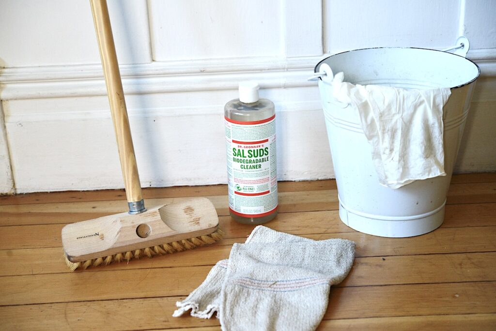 wooden scrubber brush, bottle of Sal Suds, enameled pail with rubber gloves, and big rag on wooden floor