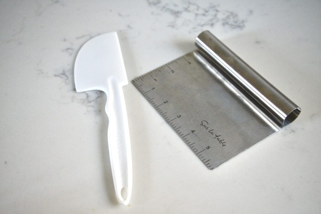 stainless steel bench scraper and white plastic bowl scraper on kitchen counter