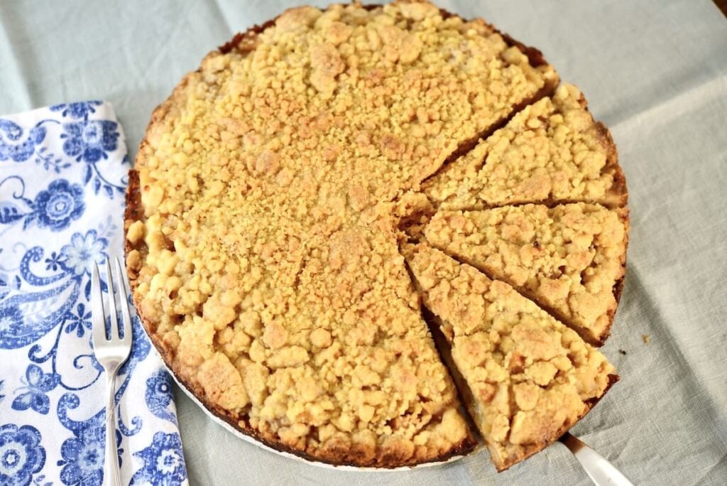 A round apple crumb cake with a crumble topping sits on a light tablecloth, several slices cut, beside a blue and white floral napkin and a fork.