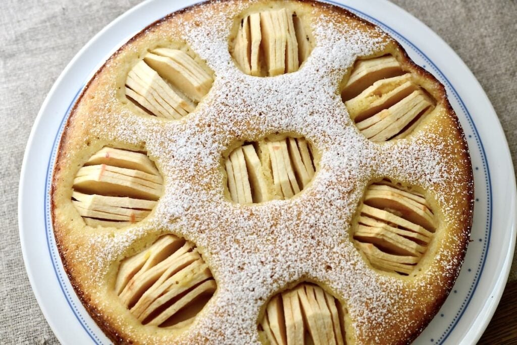 Round apple cake topped with powdered sugar, featuring several circular sections with thinly sliced apples arranged in each section, on a white plate.