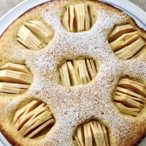 Round apple cake topped with powdered sugar, featuring several circular sections with thinly sliced apples arranged in each section, on a white plate.