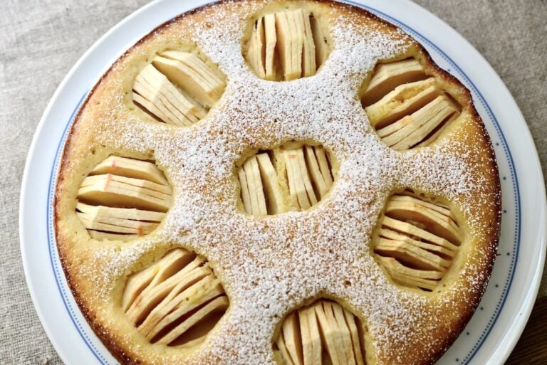 Round apple cake topped with powdered sugar, featuring several circular sections with thinly sliced apples arranged in each section, on a white plate.
