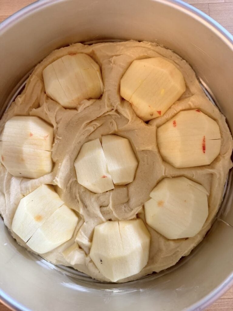 Raw cake batter in a round pan topped with six peeled, cored, and sliced apples arranged in a circle before baking.