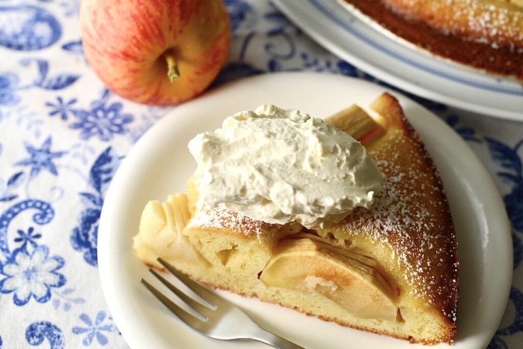 A slice of apple cake topped with whipped cream sits on a white plate with a fork. An apple and the rest of the cake are in the background on a blue-patterned tablecloth.