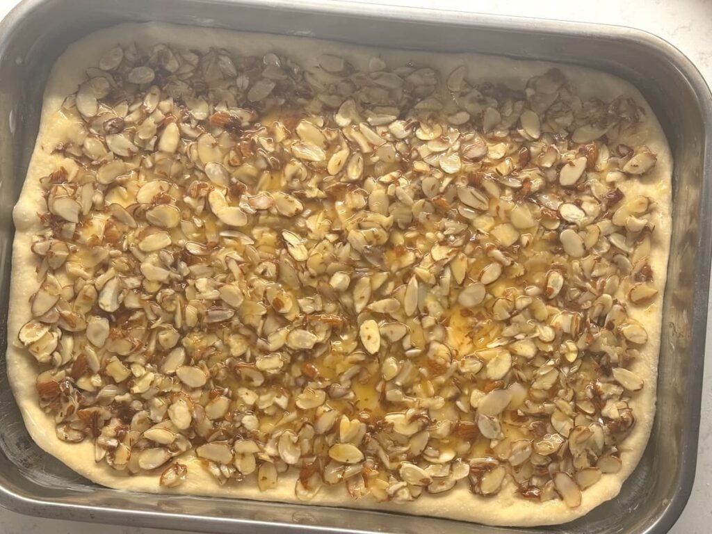 A rectangular baking pan filled with unbaked dough topped with sliced almonds and a glossy honey or syrup glaze, ready to go in the oven.