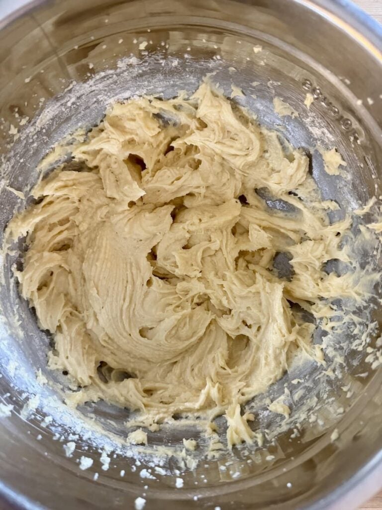 A stainless steel bowl filled with thick, creamy, pale yellow cookie dough, partially mixed, with bits of flour still visible along the edges of the bowl.