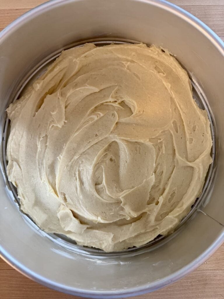 Cake batter evenly spread in a round metal baking pan, ready to be baked. The batter has a smooth, creamy texture with visible swirls from spreading.