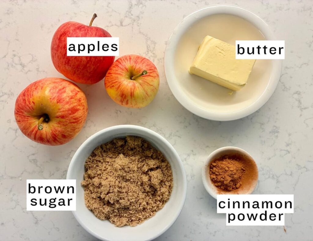 Three apples, a bowl of brown sugar, a plate with a slab of butter, and a small bowl of cinnamon powder are arranged on a countertop. Each item is labeled with text: apples, butter, brown sugar, cinnamon powder.
