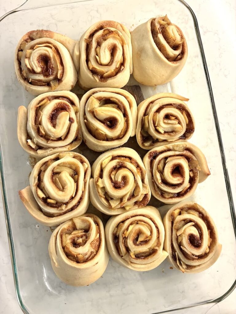 Twelve unbaked cinnamon rolls arranged closely together in a glass baking dish, ready to be baked on a light-colored countertop.