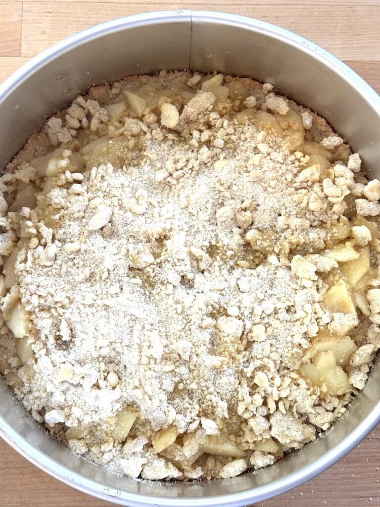 A round metal baking pan filled with an unbaked apple crumb cake, showing chopped apples and crumbly topping, rests on a wooden surface.