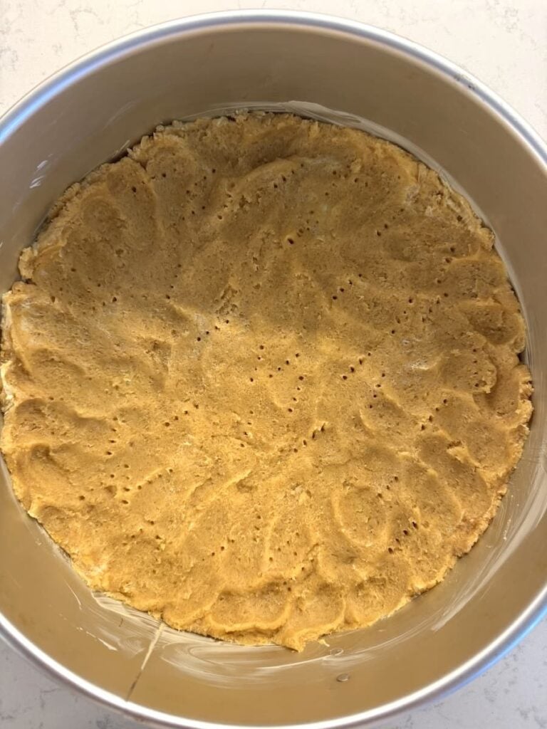 A round baking pan filled with a flattened, uncooked crust, perfect for an apple crumb cake, pressed evenly along the bottom and resembling a classic cookie or graham cracker base.