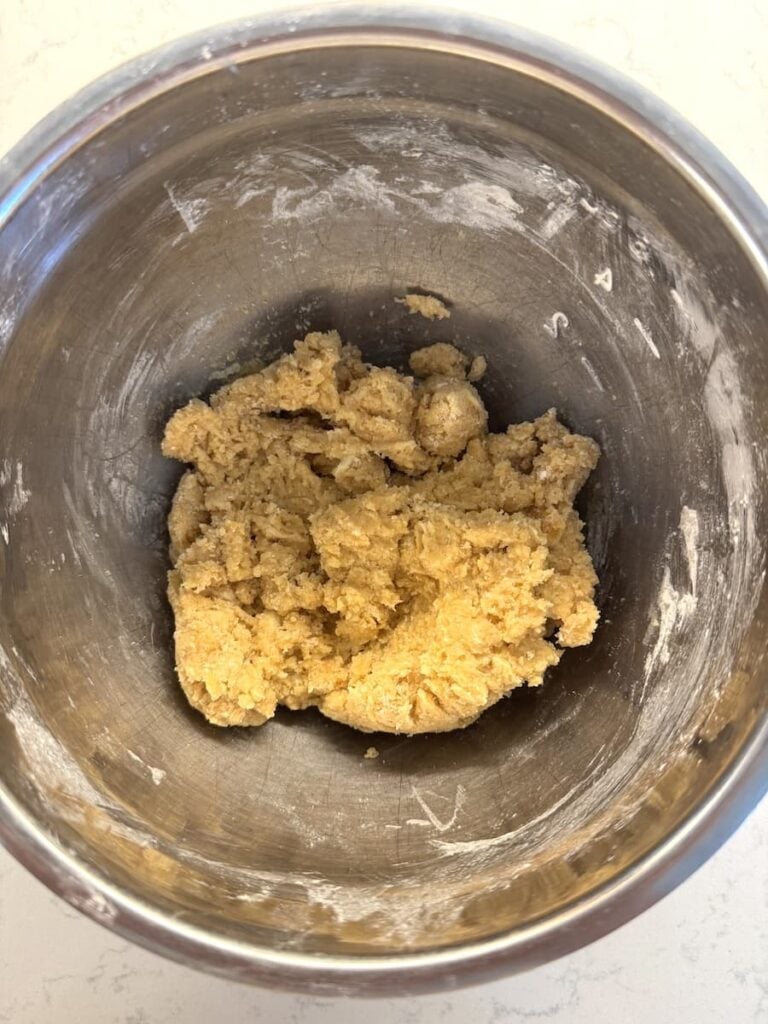 A metal mixing bowl with a ball of raw cookie dough inside, sitting on a white countertop dusted with flour—perfect for starting your next batch of apple crumb cake.