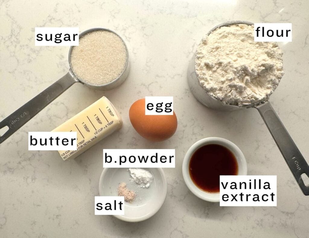 Ingredients for an apple crumb cake, including sugar, flour, butter, egg, baking powder, salt, and vanilla extract, arranged on a countertop.