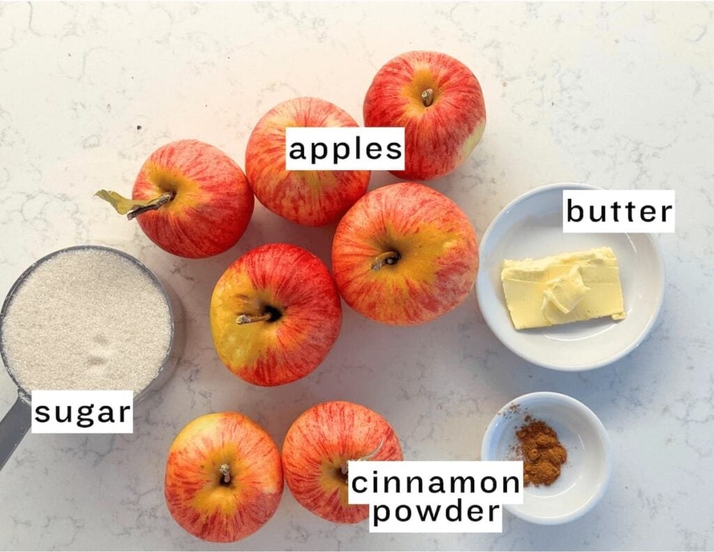 Six apples, a cup of sugar, a plate with butter, and a small bowl of cinnamon powder are arranged on a light countertop—perfect ingredients for an apple crumb cake. Each ingredient is labeled with text.