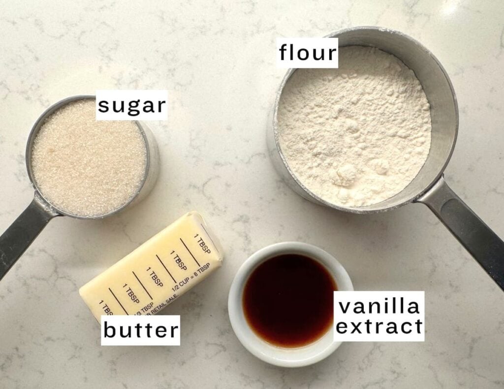 Measuring cups of sugar and flour, a stick of butter, and a small bowl of vanilla extract for an apple crumb cake, all labeled with their names.