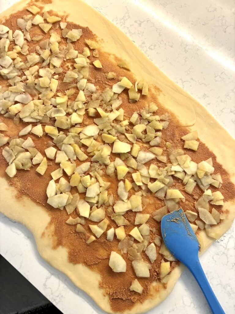 Rectangular rolled out dough topped with cinnamon, sugar, and chopped apples, with a blue spatula resting on one corner on a light-colored countertop.