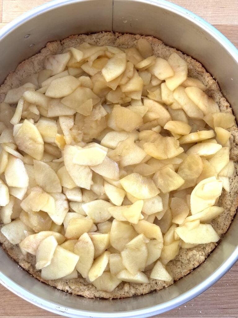 A round baking pan filled with a layer of sliced apples on top of a crust, ready for baking, sits on a wooden surface—perfect for an apple crumb cake.