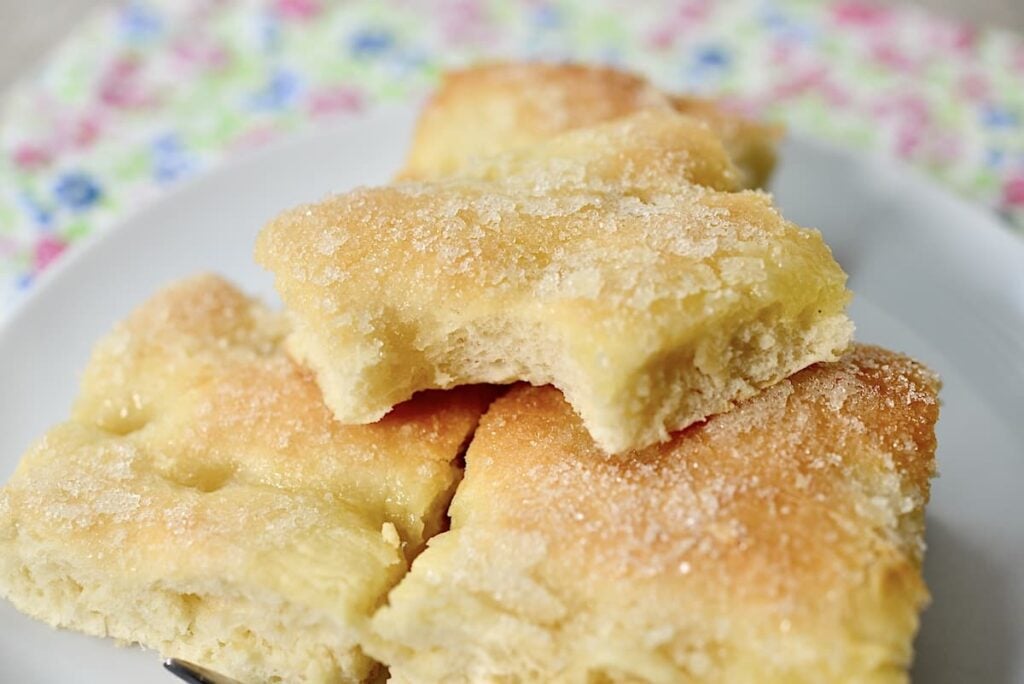 Four pieces of sugar-dusted focaccia bread, reminiscent of German butterkuchen, are stacked on a white plate, with a floral-patterned tablecloth visible in the background. The bread looks soft and slightly golden.