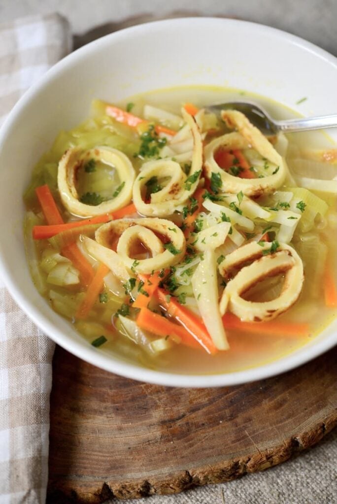A bowl of clear German pancake soup with julienned carrots, cabbage, and golden-brown spiral egg noodles, garnished with chopped herbs. A spoon rests in the bowl, which sits on a wooden surface next to a checkered cloth.
