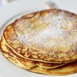 A stack of golden-brown sourdough pancakes dusted with powdered sugar sits on a white plate, with a fork partially visible in the background.