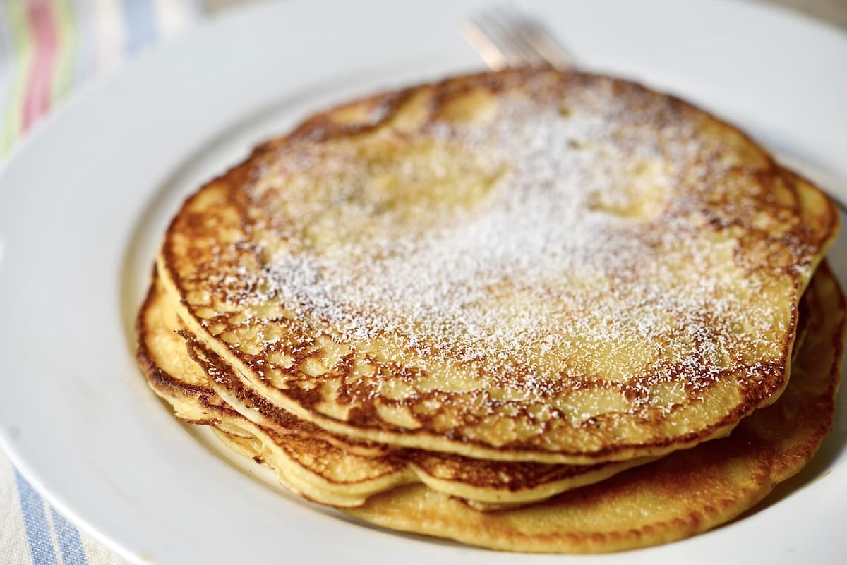 A stack of golden-brown sourdough pancakes dusted with powdered sugar sits on a white plate, with a fork partially visible in the background.