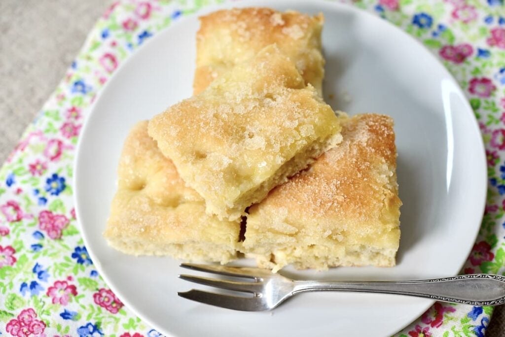 Three pieces of sugar-topped German butterkuchen focaccia bread are stacked on a white plate with a small fork, resting on a colorful floral-patterned cloth.