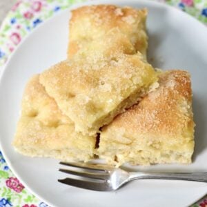 Three pieces of sugar-topped German butterkuchen focaccia bread are stacked on a white plate with a small fork, resting on a colorful floral-patterned cloth.
