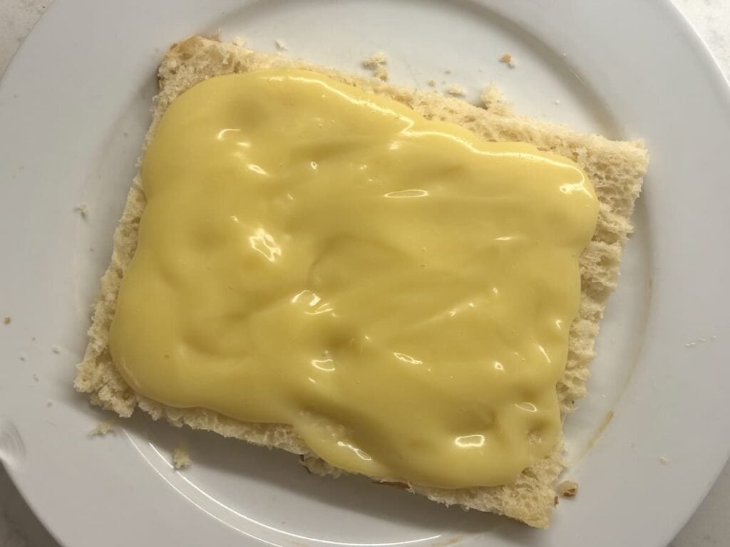 A slice of white bread on a white plate, topped with a thick layer of yellow custard or pudding.