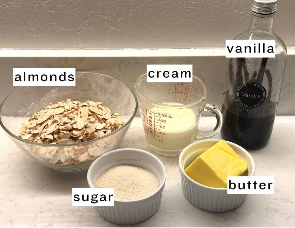 A countertop displays labeled ingredients in bowls and containers: sliced almonds, a measuring cup of cream, a bottle of vanilla extract, a small bowl of sugar, and a small bowl of butter.