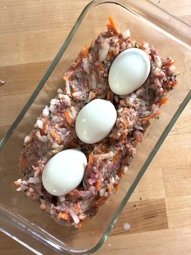 A rectangular glass dish filled with raw ground meat mixture topped with three peeled hard-boiled eggs.