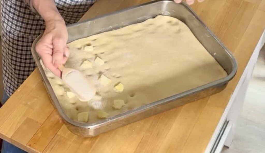 A person prepares German butterkuchen by sprinkling small cubes of butter onto dough spread in a rectangular metal baking tray on a wooden countertop.
