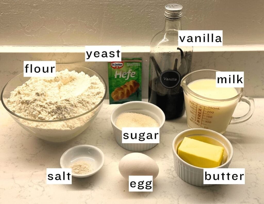 A countertop displays labeled baking ingredients for German butterkuchen: a bowl of flour, a packet of yeast, a bottle of vanilla, a measuring cup of milk, sugar, butter, salt, and a whole egg.